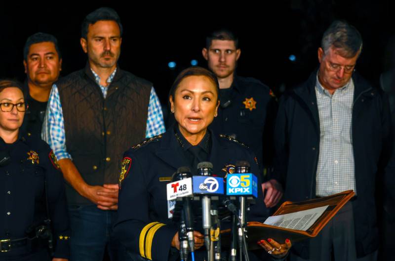 A small Latina woman stands at a podium in front of multiple microphones, with people standing behind her.