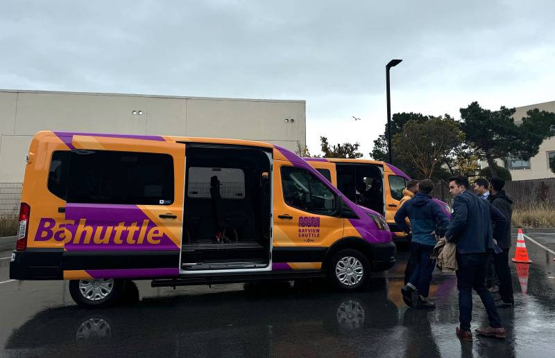 A small group of people stand outside next to two purple and yellow vans that say 'Bayview Community Shuttle.'