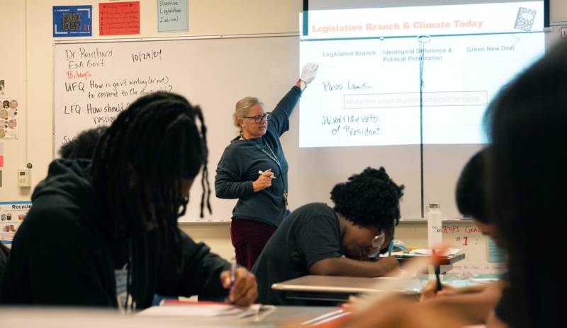 A white woman wearing glasses and a navy blue shirt points at whiteboard with a projected image as students sit at their desks and work.