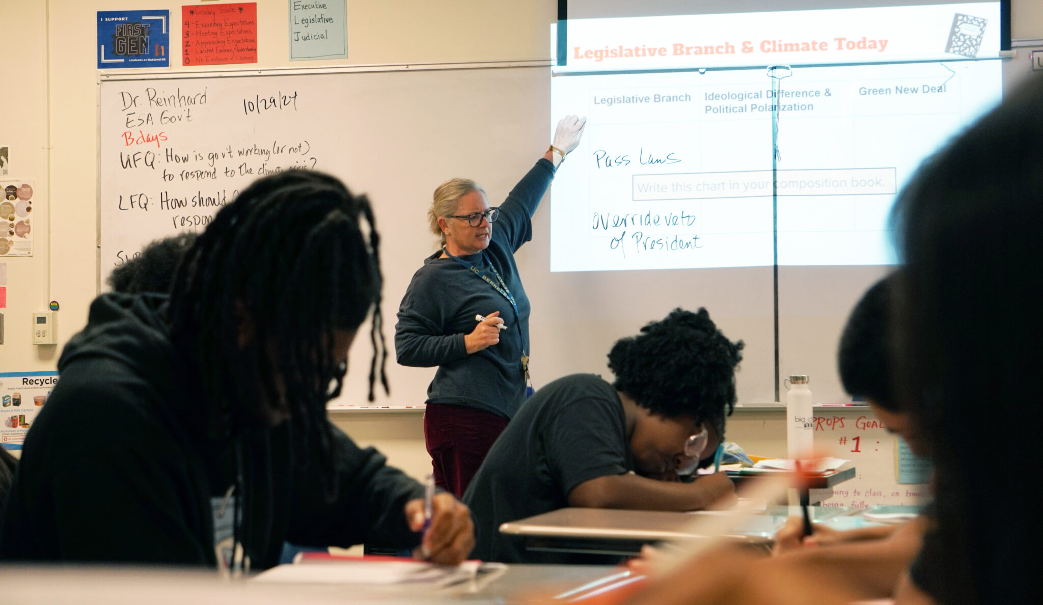 A white woman wearing glasses and a navy blue shirt points at whiteboard with a projected image as students sit at their desks and work.