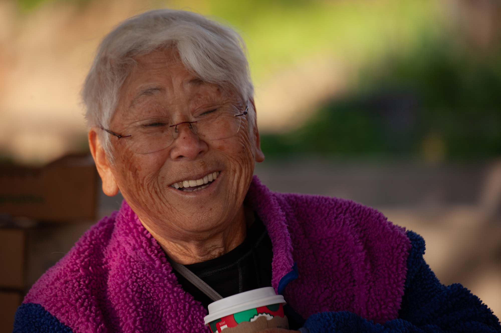 A person with white hair and glasses, smiles while wearing a maroon top. 