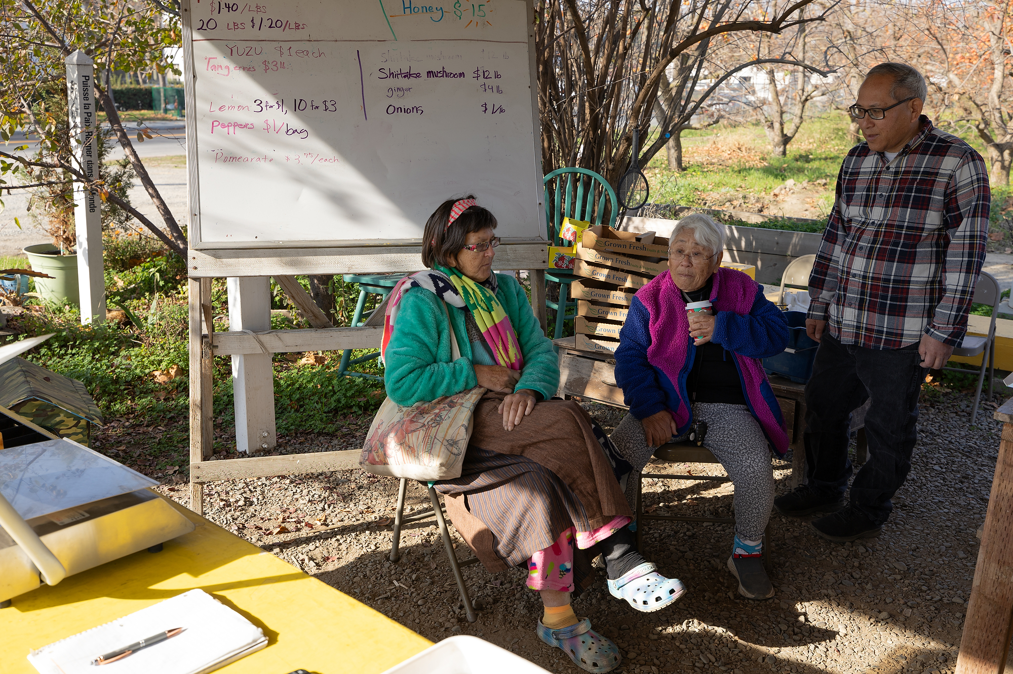 Two people sit in sun and shade at a farm stand with a white board behind them.