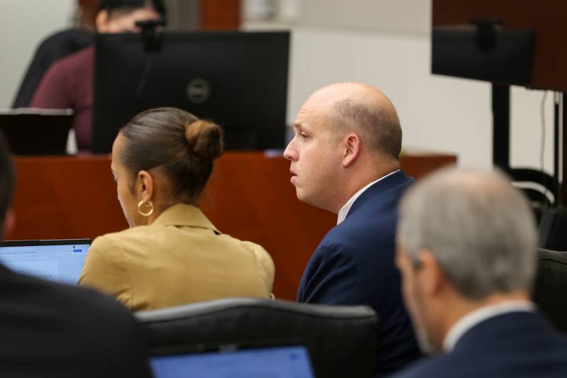 A bald man sits in a courtroom next to a woman.