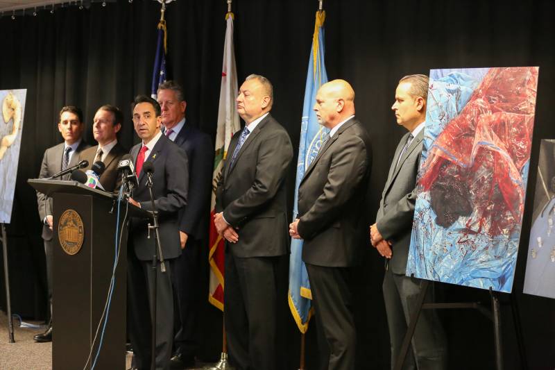 Men in suits stand behind a podium, next to a bloody photo.