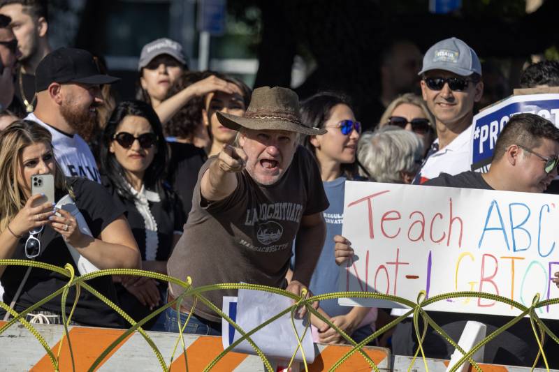 A man in a brown hat and brown shirt points aggressively with several people in the background.