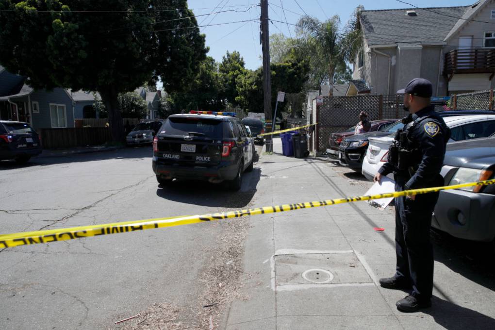 A police officer stands behind yellow caution tape on a city street, with a police car in the background.