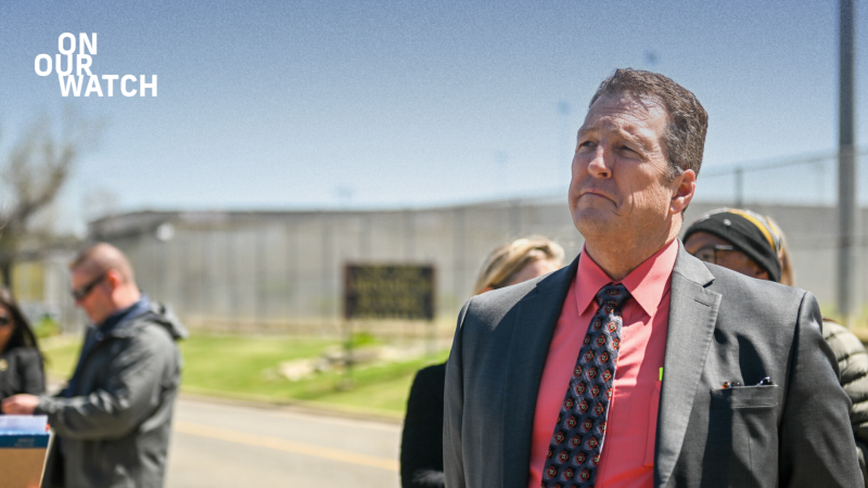 The warden of New Folsom Prison, a broad-shouldered, middle-aged man with short brown hair, stands in front of a road during a press tour. He is wearing a bright salmon-colored shirt and a dark suit with a patterned tie, looking into the distance with a serious expression. The prison’s chain link fence is visible in the background, along with other attendees, some taking notes. The ‘On Our Watch’ logo is in the top-left corner.