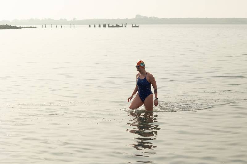 A woman in a bathing suit and swim cap wades out into a vast body of water.