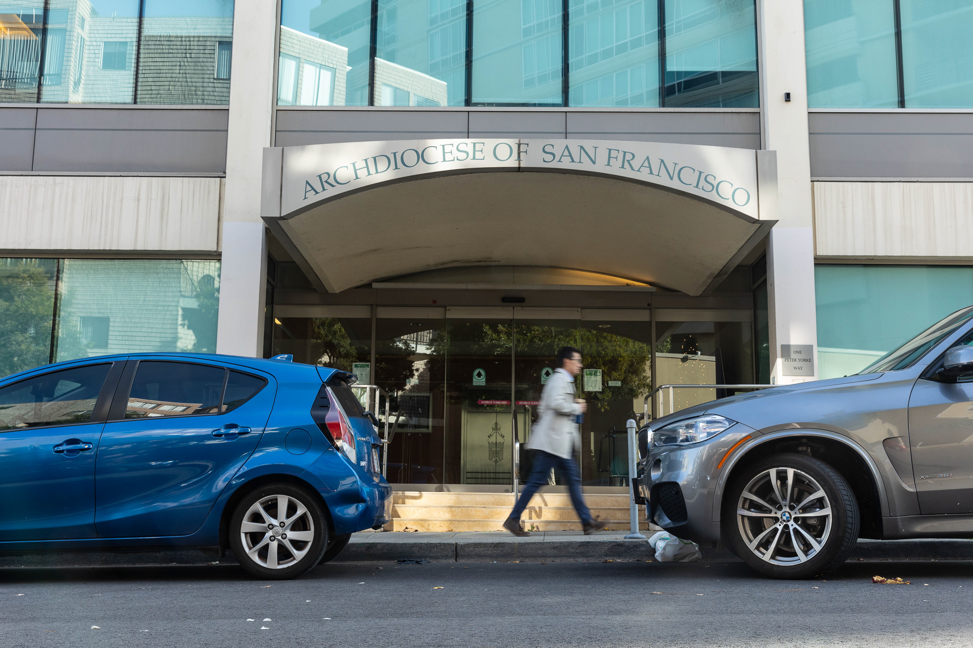 A person walks past two cars and a building with a sign above them that says "Archdiocese of San Francisco."