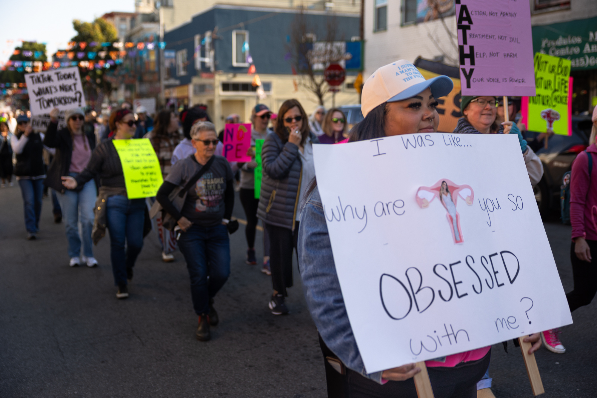 A woman wearing a white hat holds a white sign with the word "Obsessed" in large letters.