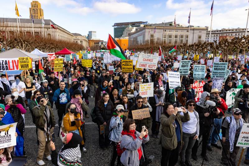 Several people in a large crowd hold signs and flags with buildings in the background.