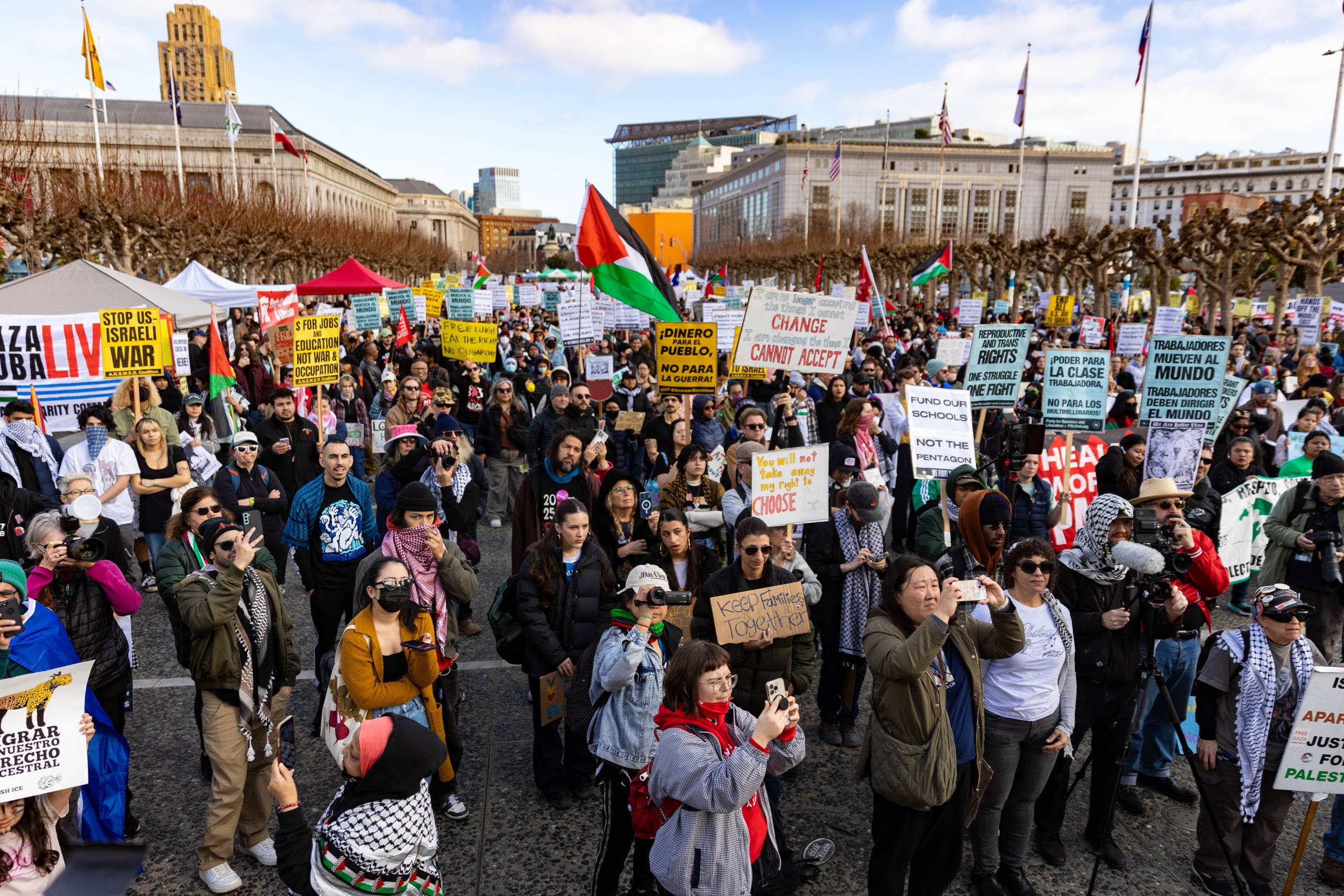 Several people in a large crowd hold signs and flags with buildings in the background.