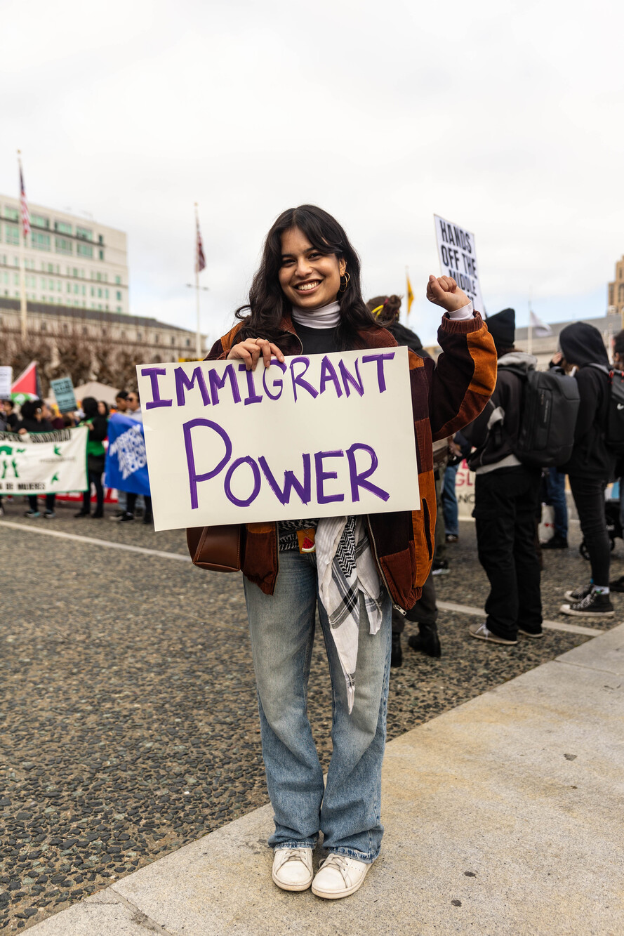 A woman holds a sign that says "Immigrant Power" near a crowd.