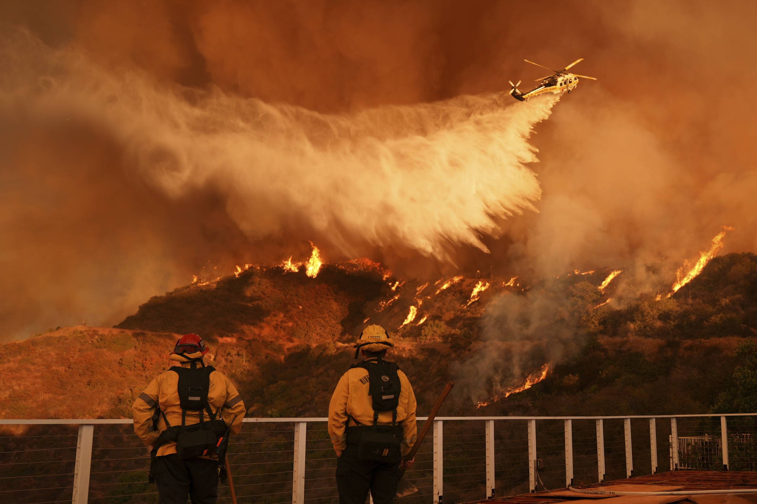 Plane drops water as firefighters look on with smoke and flames below.