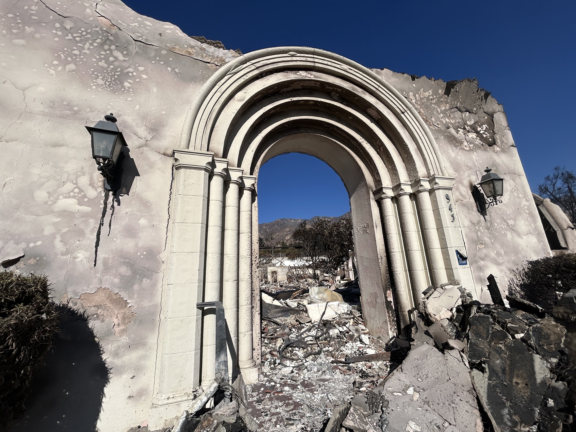 A front archway to a tan church stands amongst the charred rubble of a burned church.