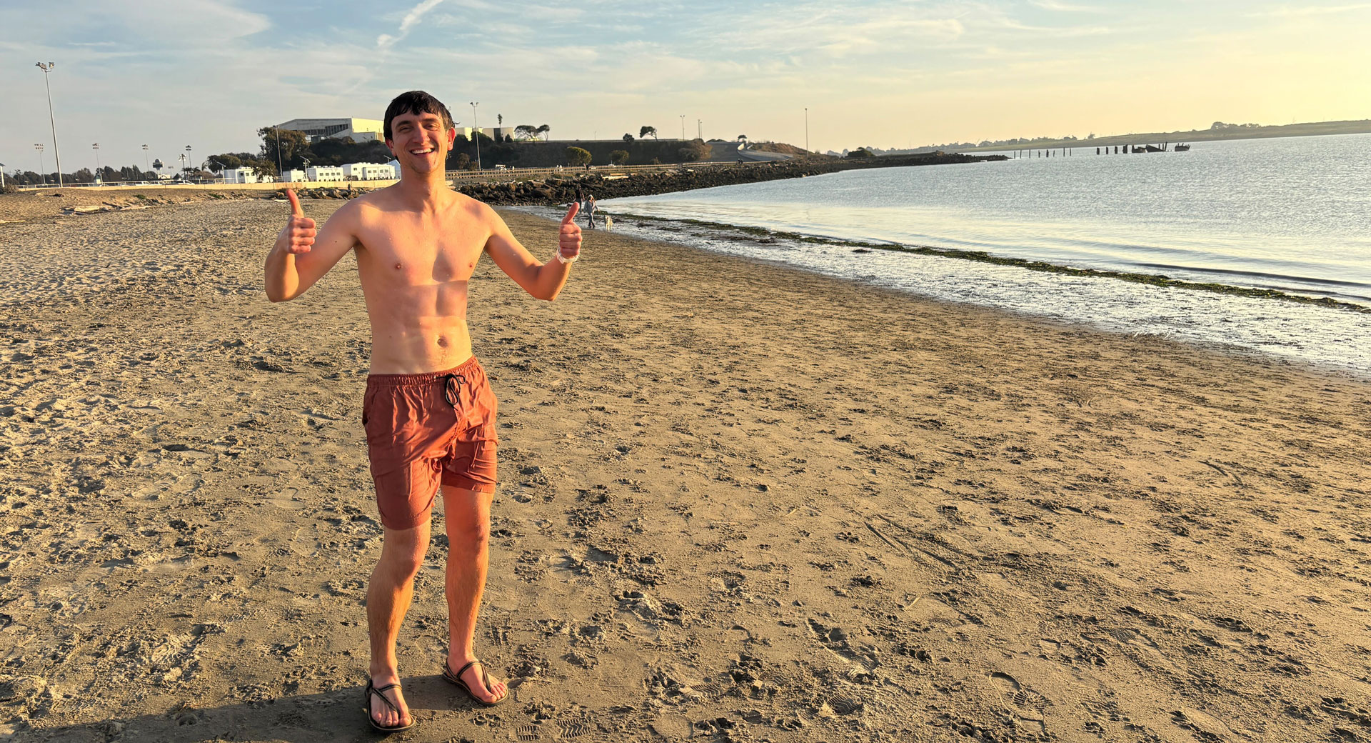 A man gives the thumbs up sign standing on a beach next to a large body of water.