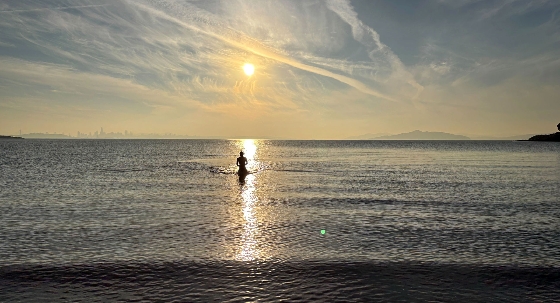 The silhouette of man standing out in a large body of water, looking at the sunset behind a cityscape.