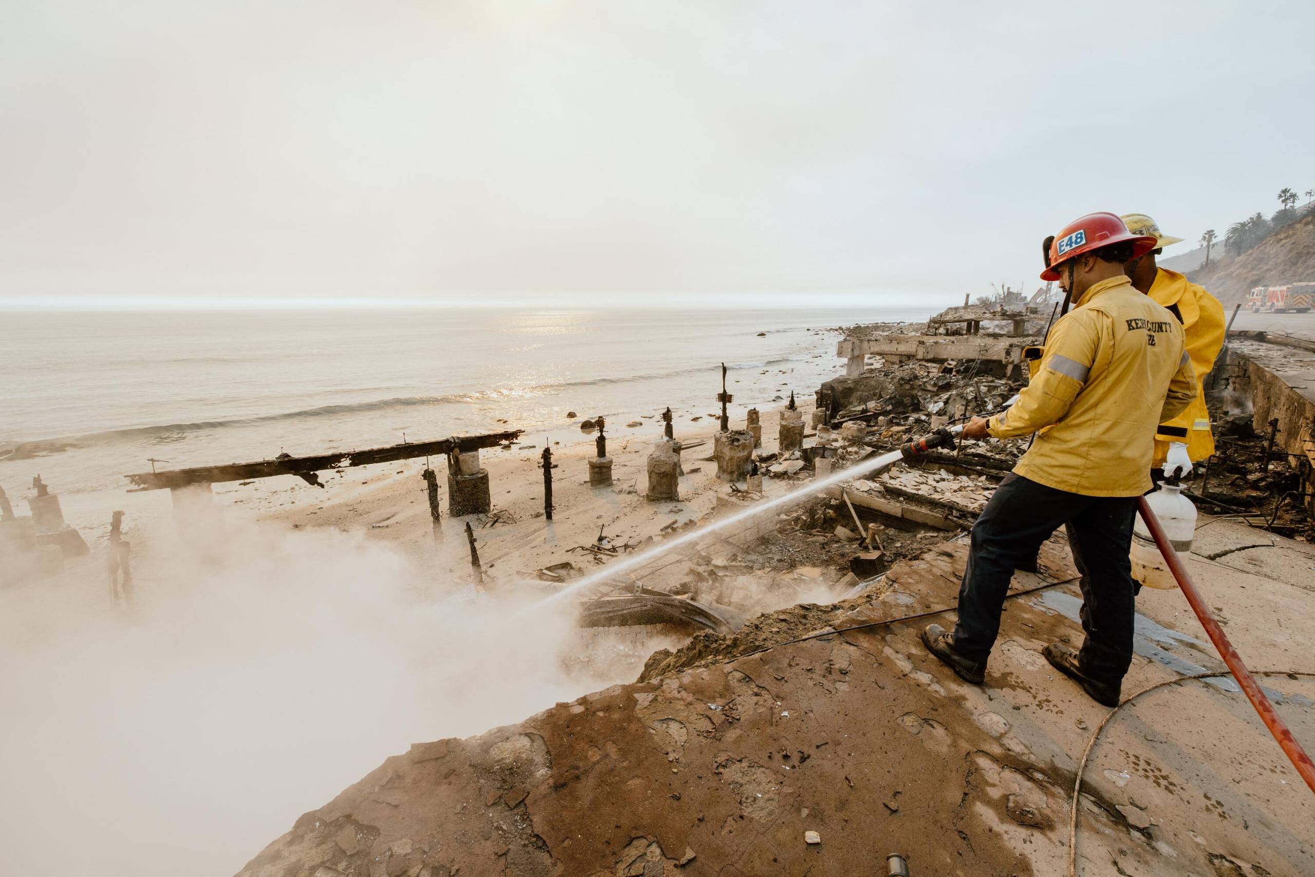 Two men dressed in firefighter uniforms with red helmets hold a fire hose