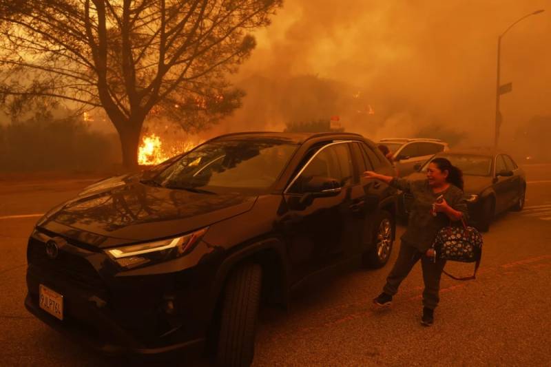 A woman cries by a car as she tries to get in with orange glowing smoke behind her on a fire-engulfed street.