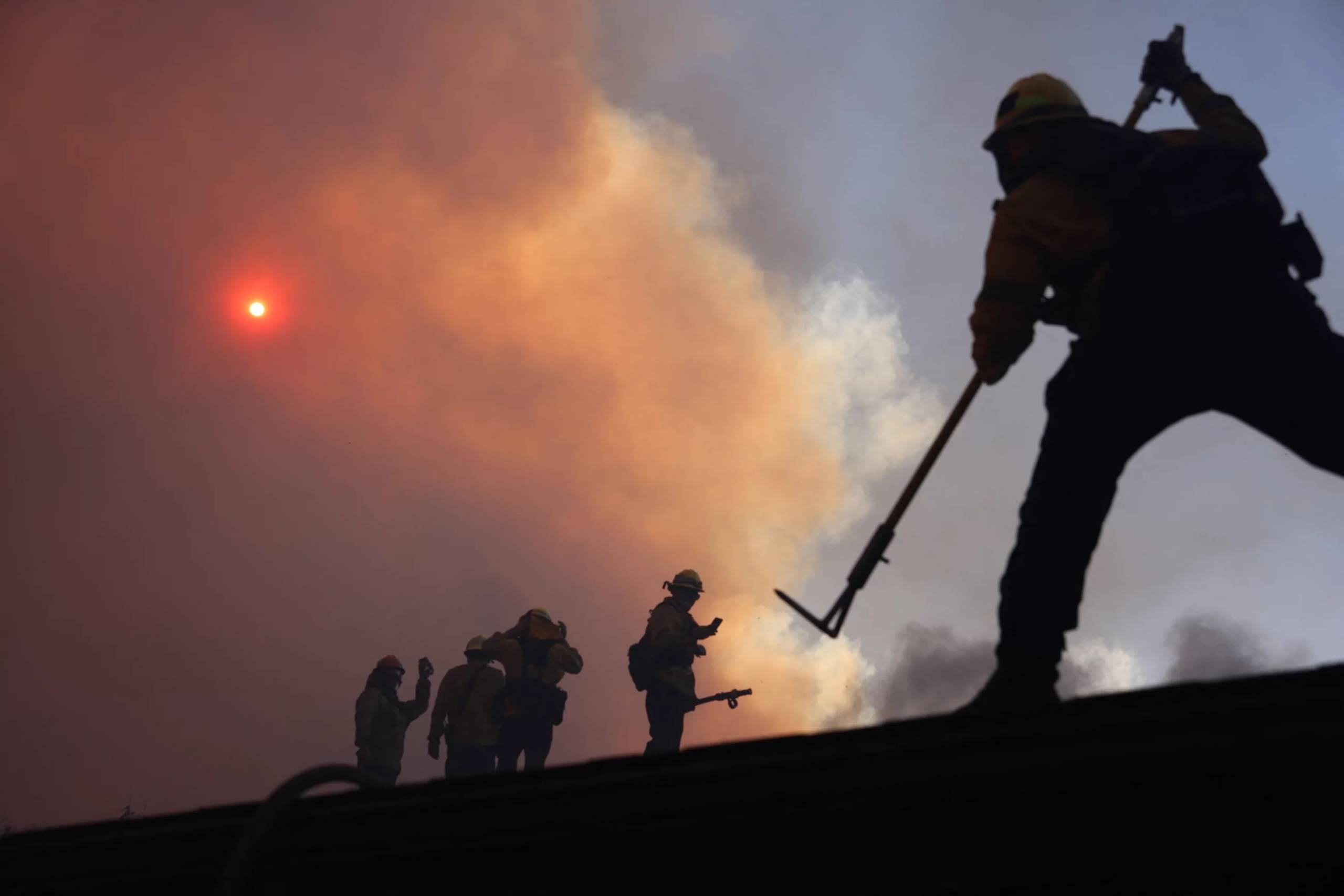 A plume of smoke covers the sun glowing orange above firefighters working with tools to contain the fire.