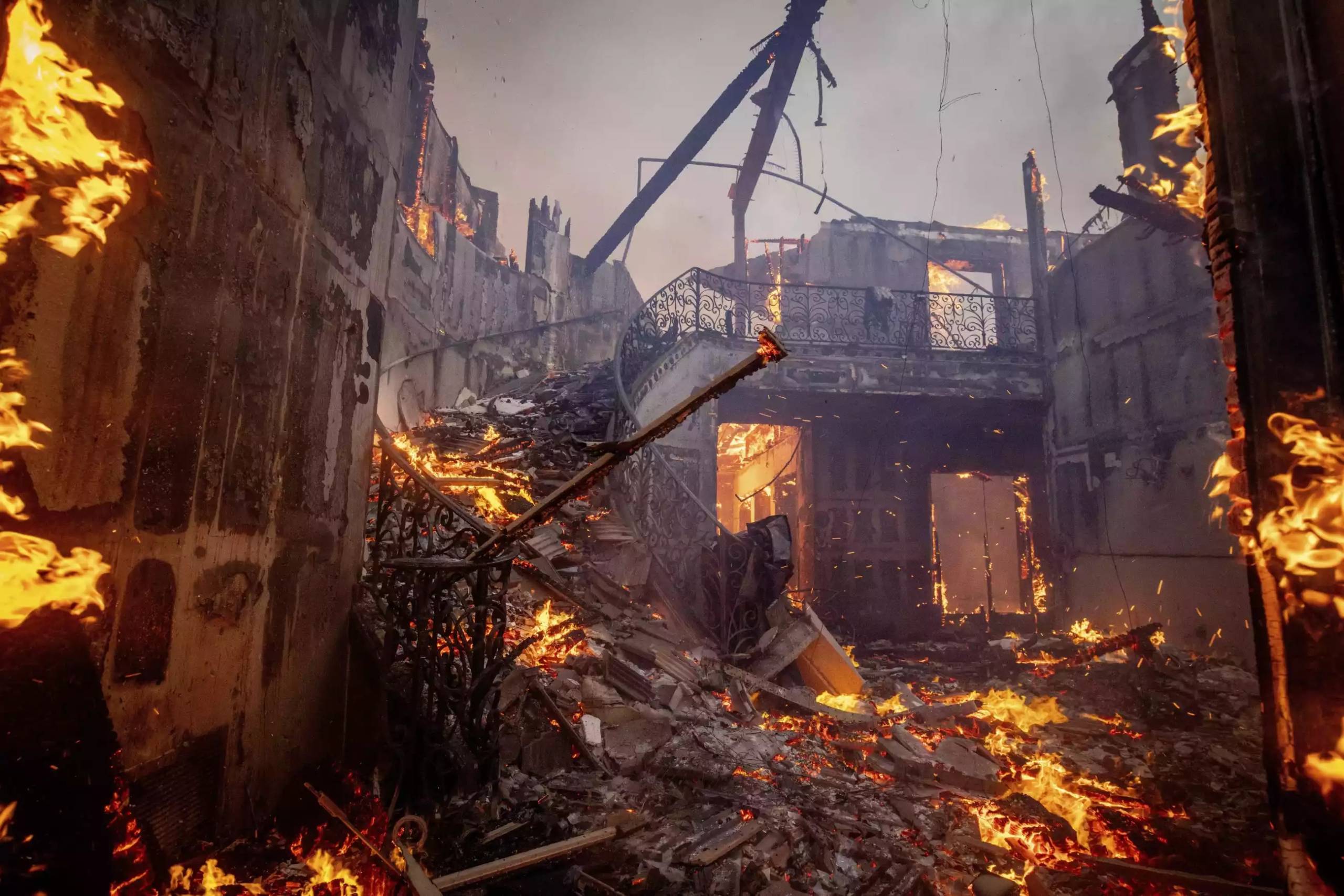 The wreckage of the foyer of a house with the second floor completely collapsed and a stairway still on fire.