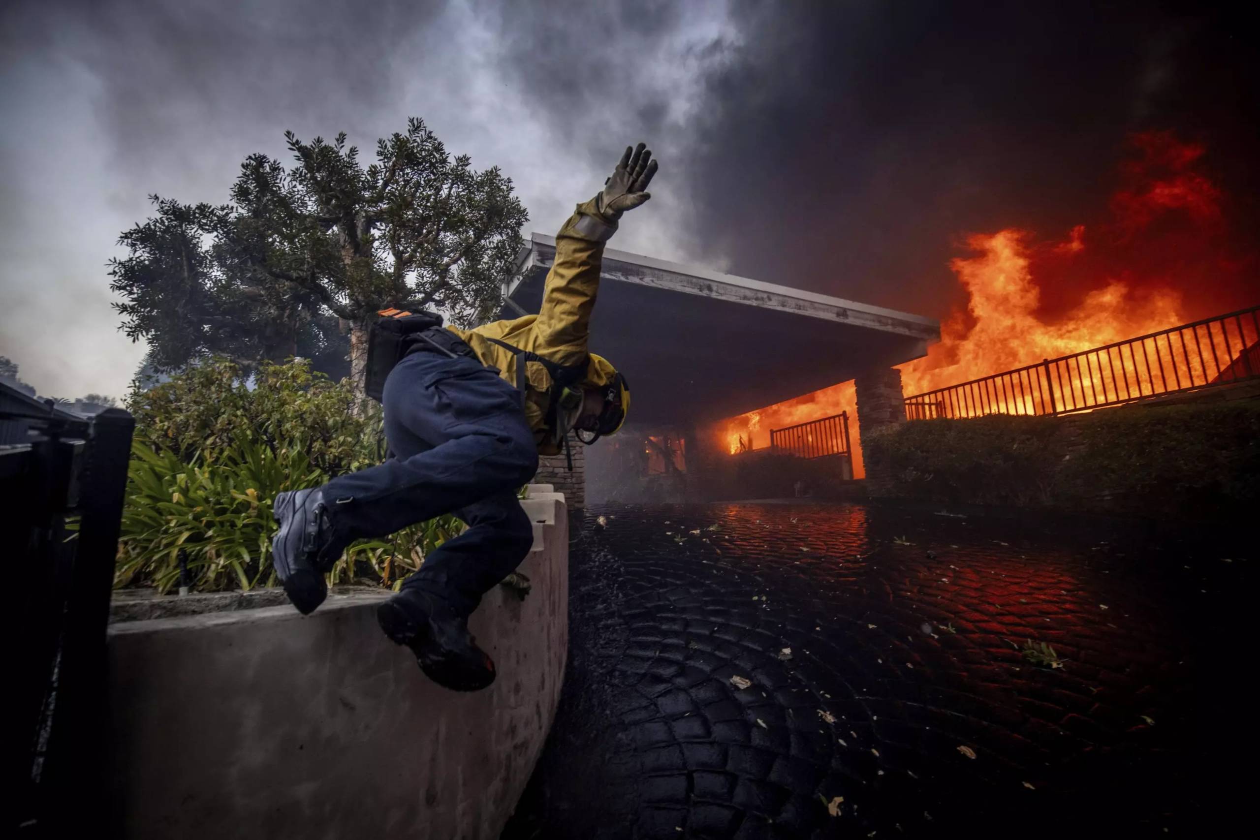 A firefighter jumps over a low wall and toward a burning house.