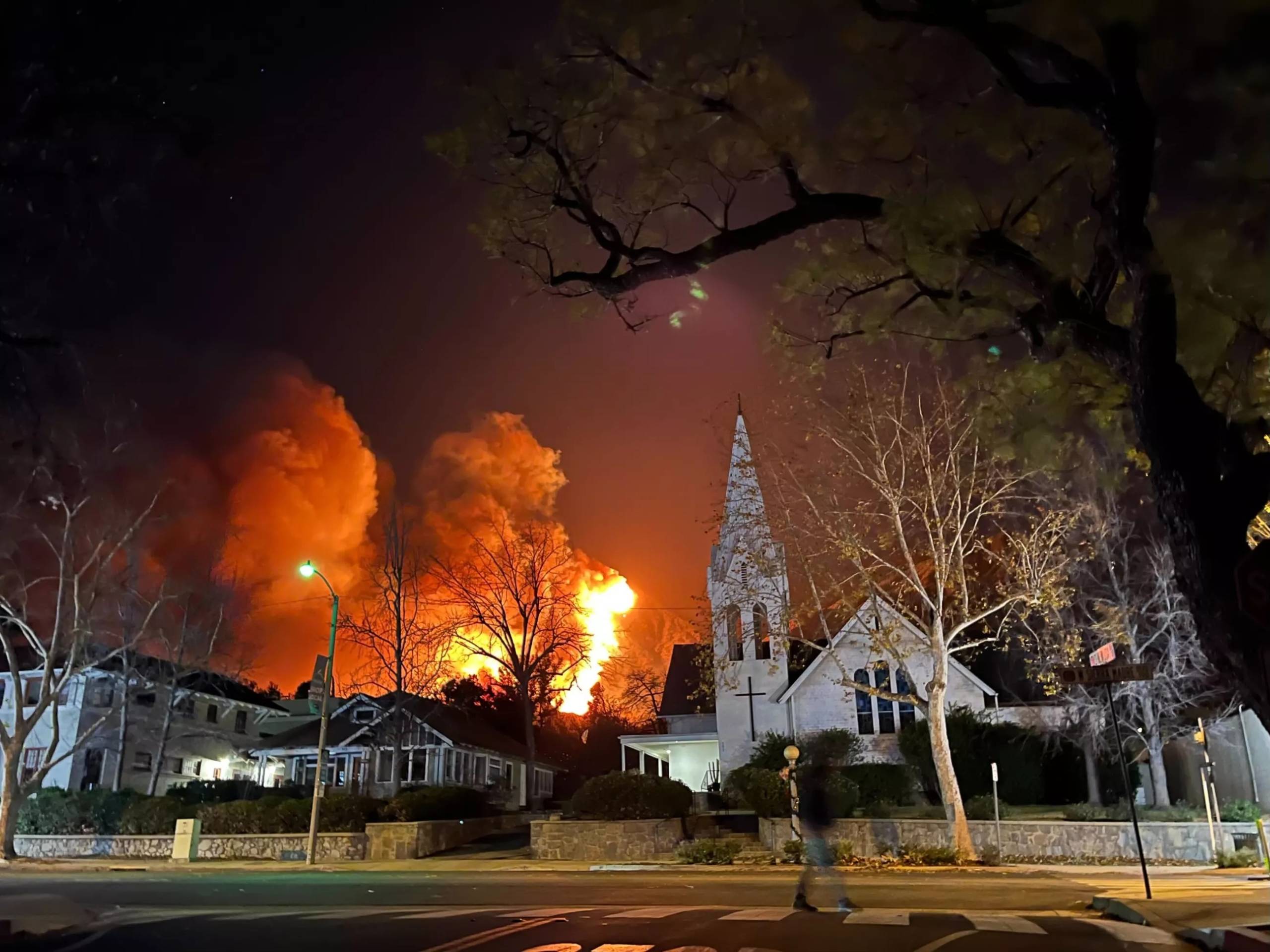 A city street with houses and a church, flames rising behind them, and a person walking in the street in the foreground.