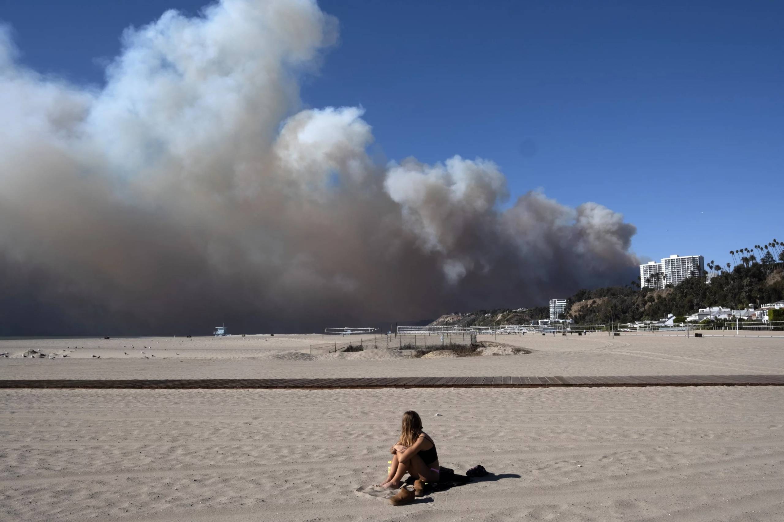 A lone woman on a beach looks over her shoulder at a massive plume of smoke in the near distance.