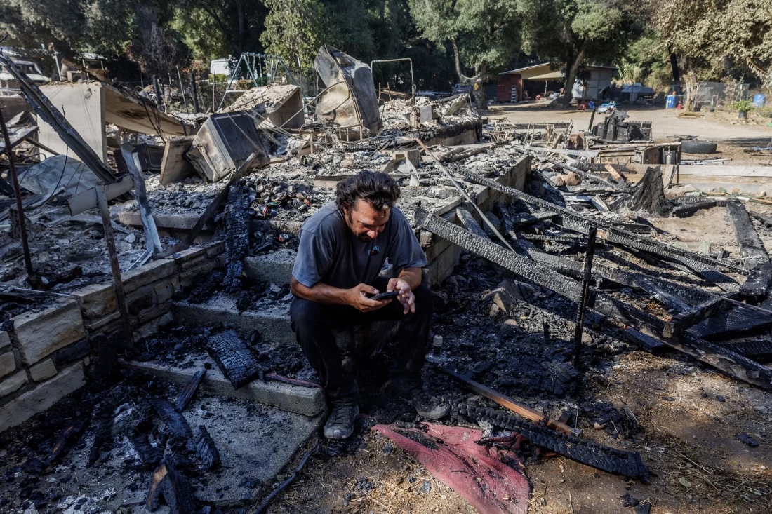 A man sits amid ruins of a house as he looks at and tries to operate his smartphone.