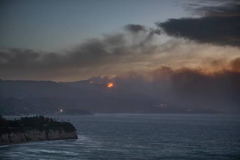 Fire and flame with smoke rising on a distant wooded mountain with the sea in the foreground.