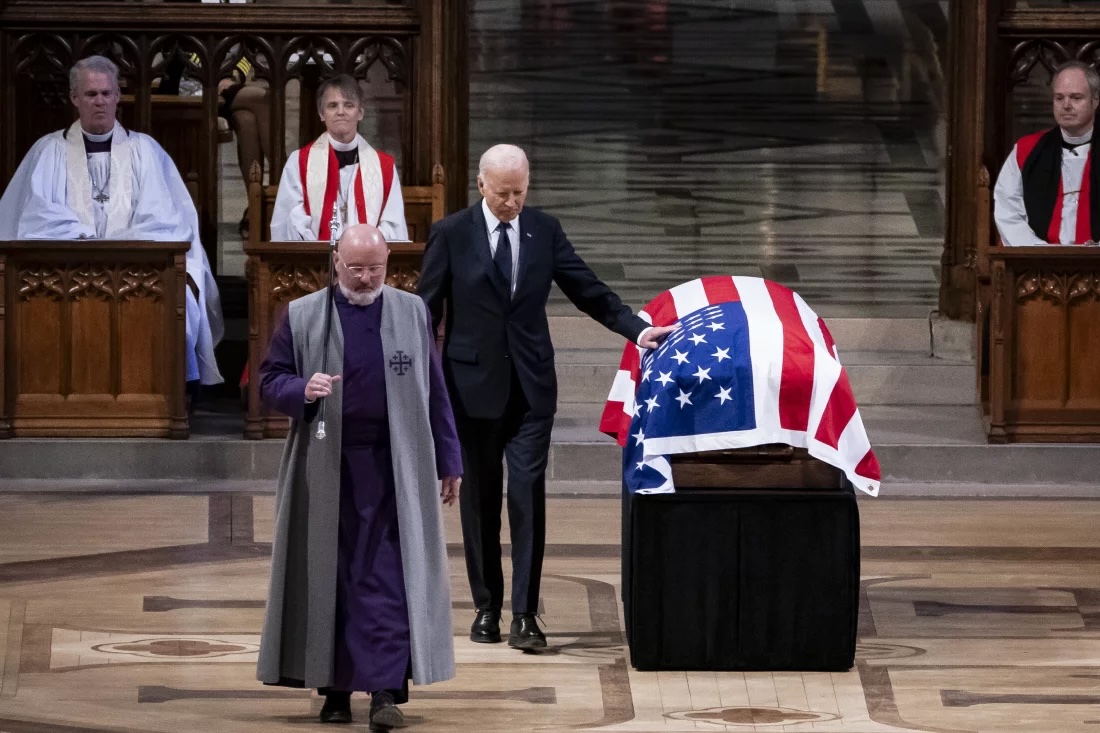 An older man in a suit and tie lays a hand on a casket with a U.S. flag draped over it with a priest before him and clergymen behind him.