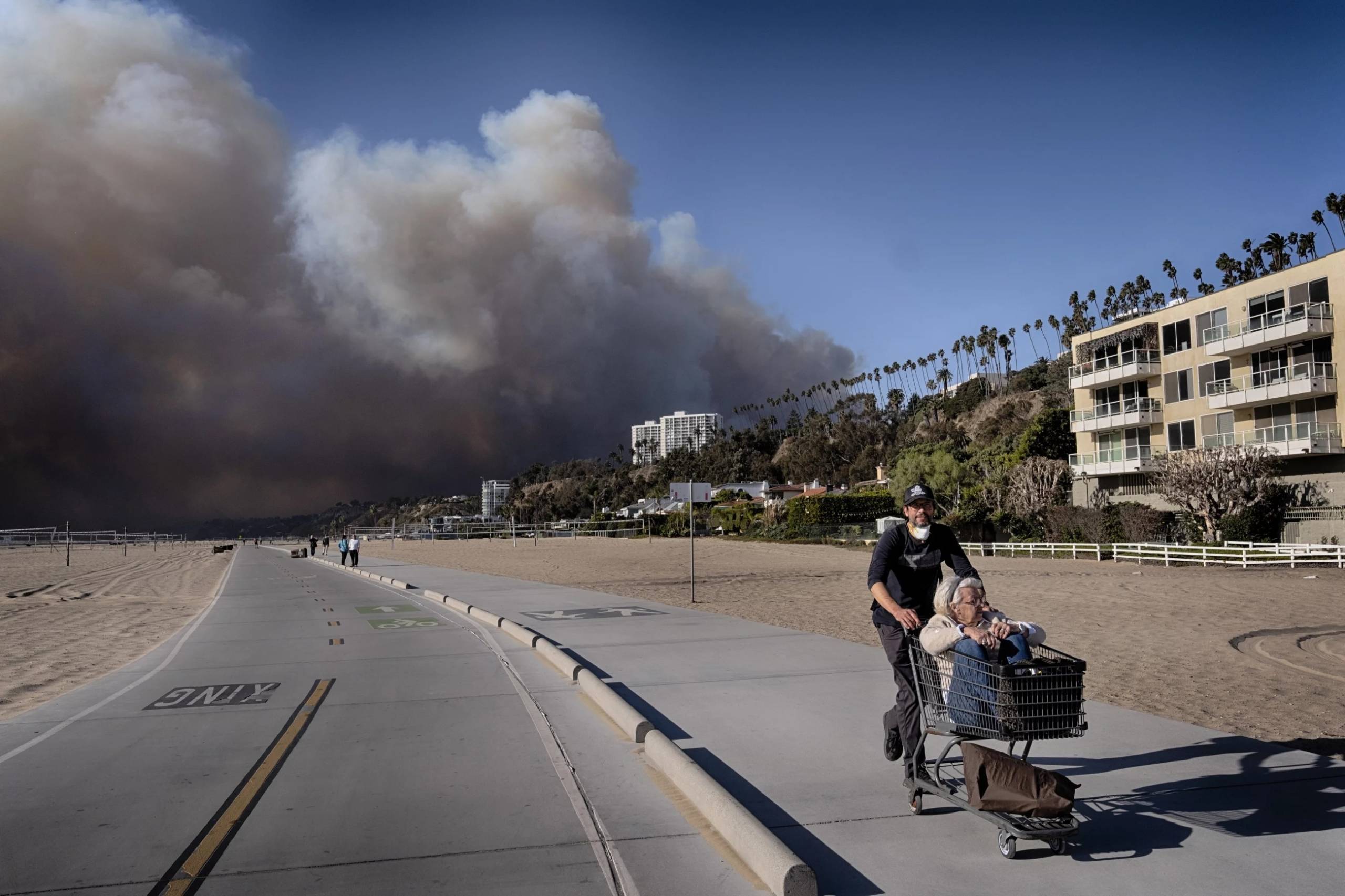 Man pushes an older woman sitting in a shopping cart away from a plume of smoke along the sidewalk of a beach.