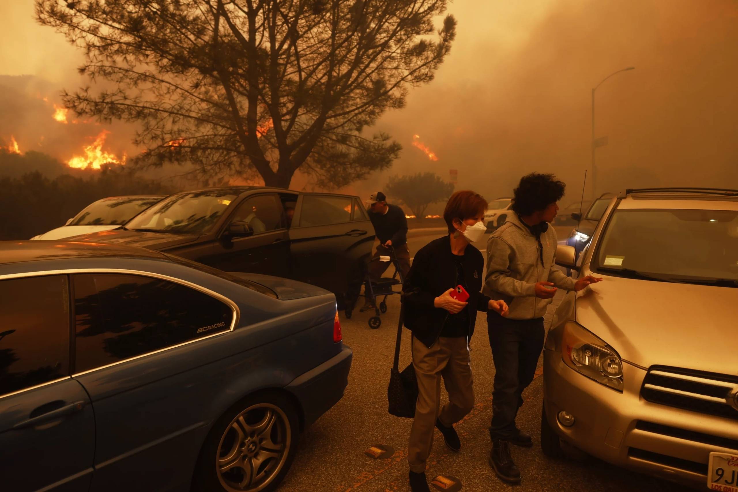 People fleeing among cars with a tree and orange smoky haze and flames in the background.