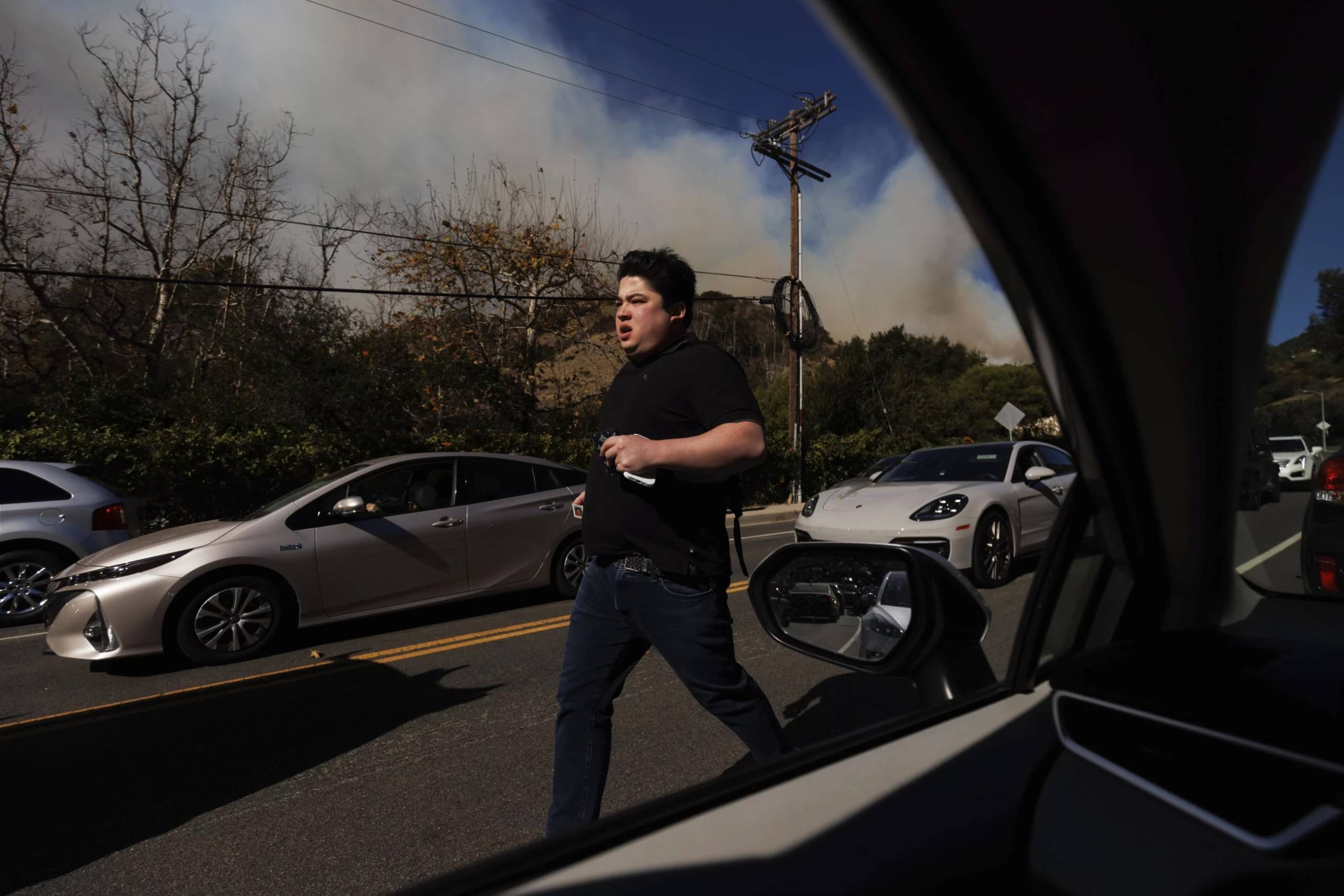 A young man runs with a worries face past cars on a city street with smoke in the distance.