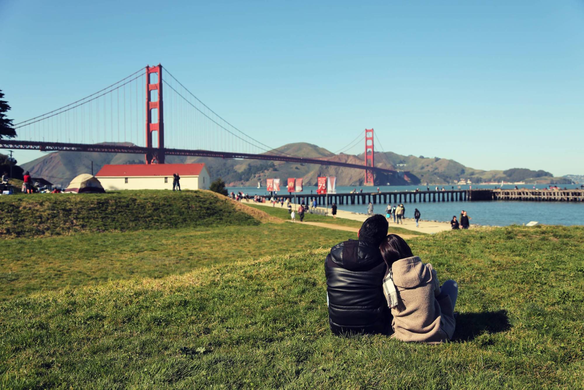 A couple leans on each other as they sit on the grass and overlook the Golden Gate Bridge.