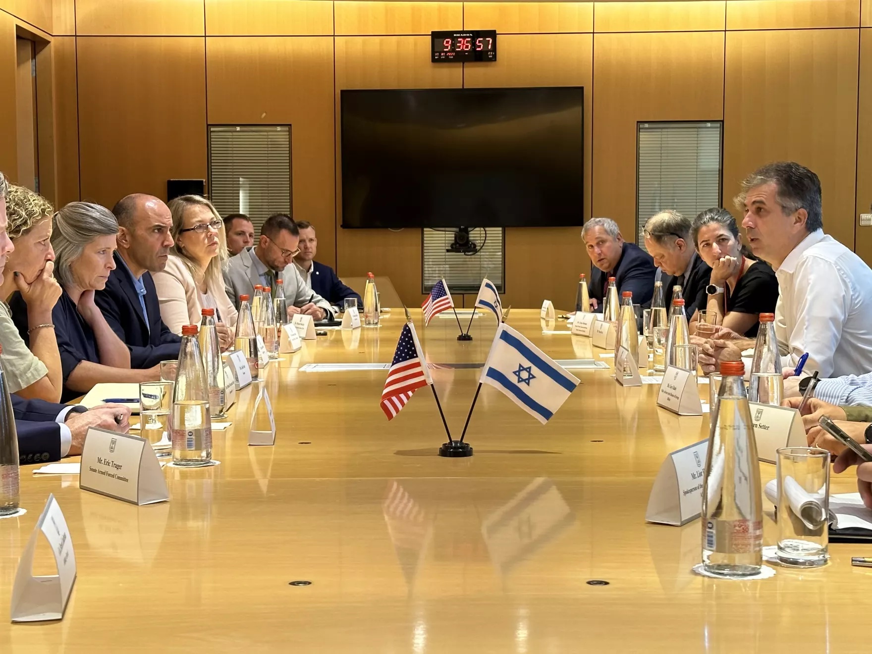 A large group of people sit around a big table with small U.S. and Israeli flags in the middle.