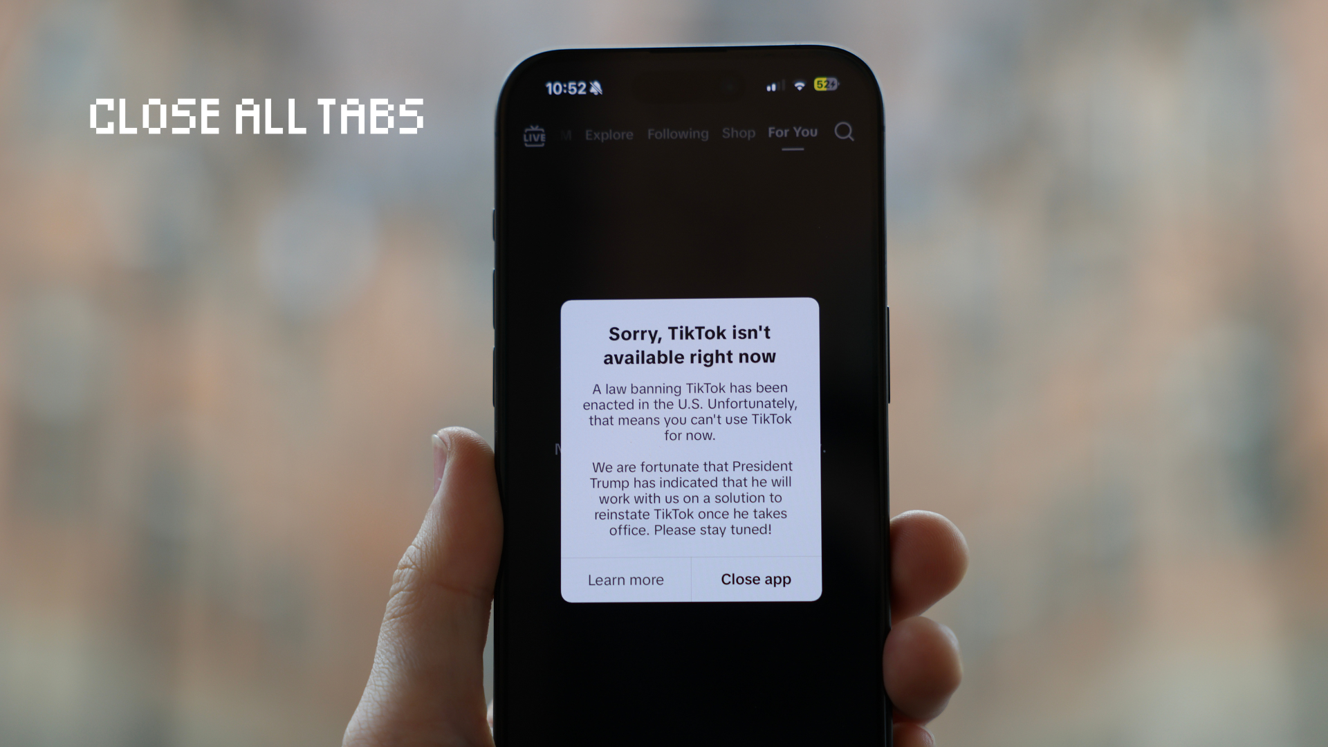 A hand holds an iPhone, which displays a popup message that reads "Sorry, TikTok isn't available right now." The background is blurred.