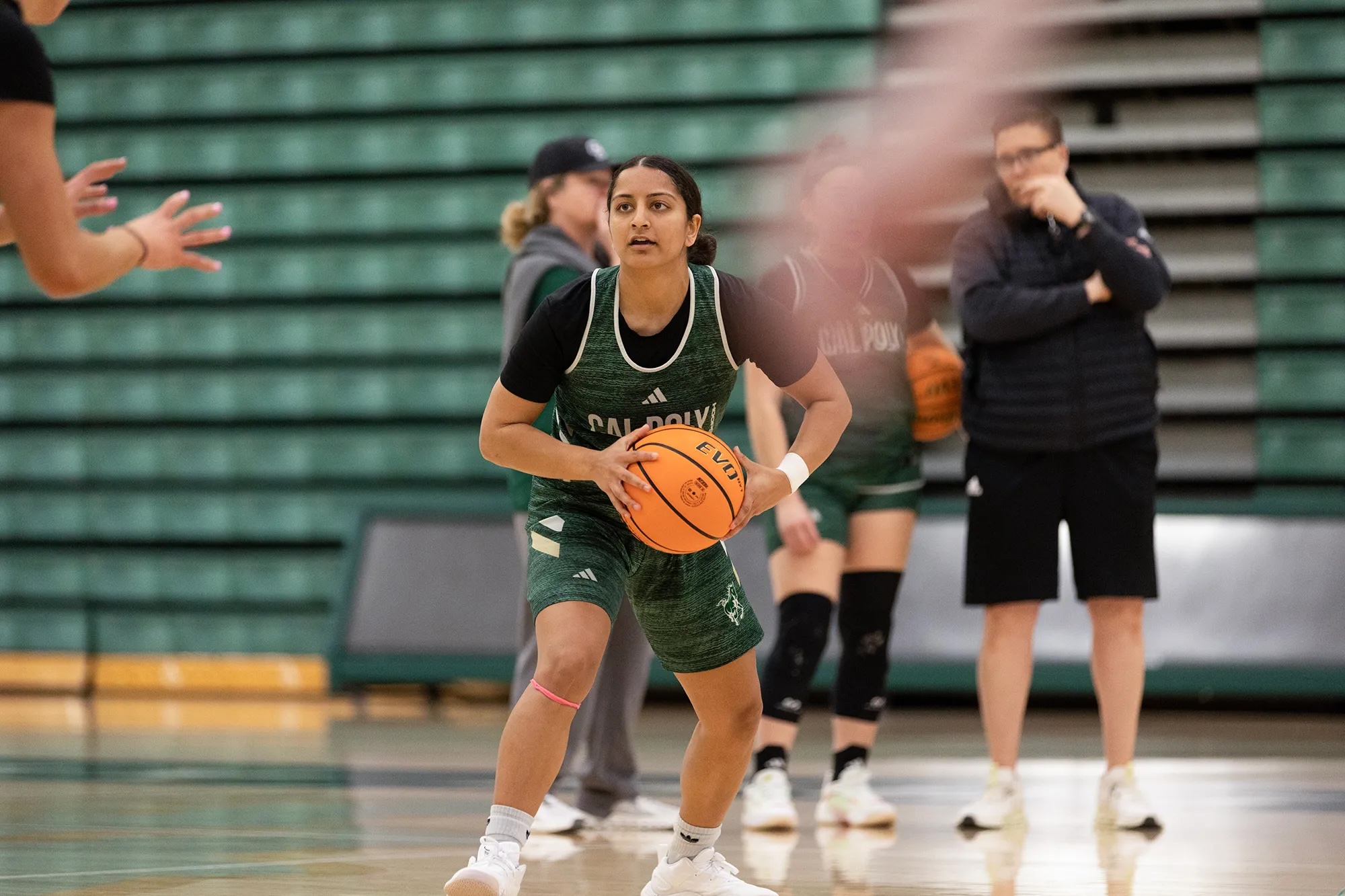 A female basketballer looks at the hoop, ready for a jump shot, she's wearing a green outfit with empty bleachers behind her and other players in the background.