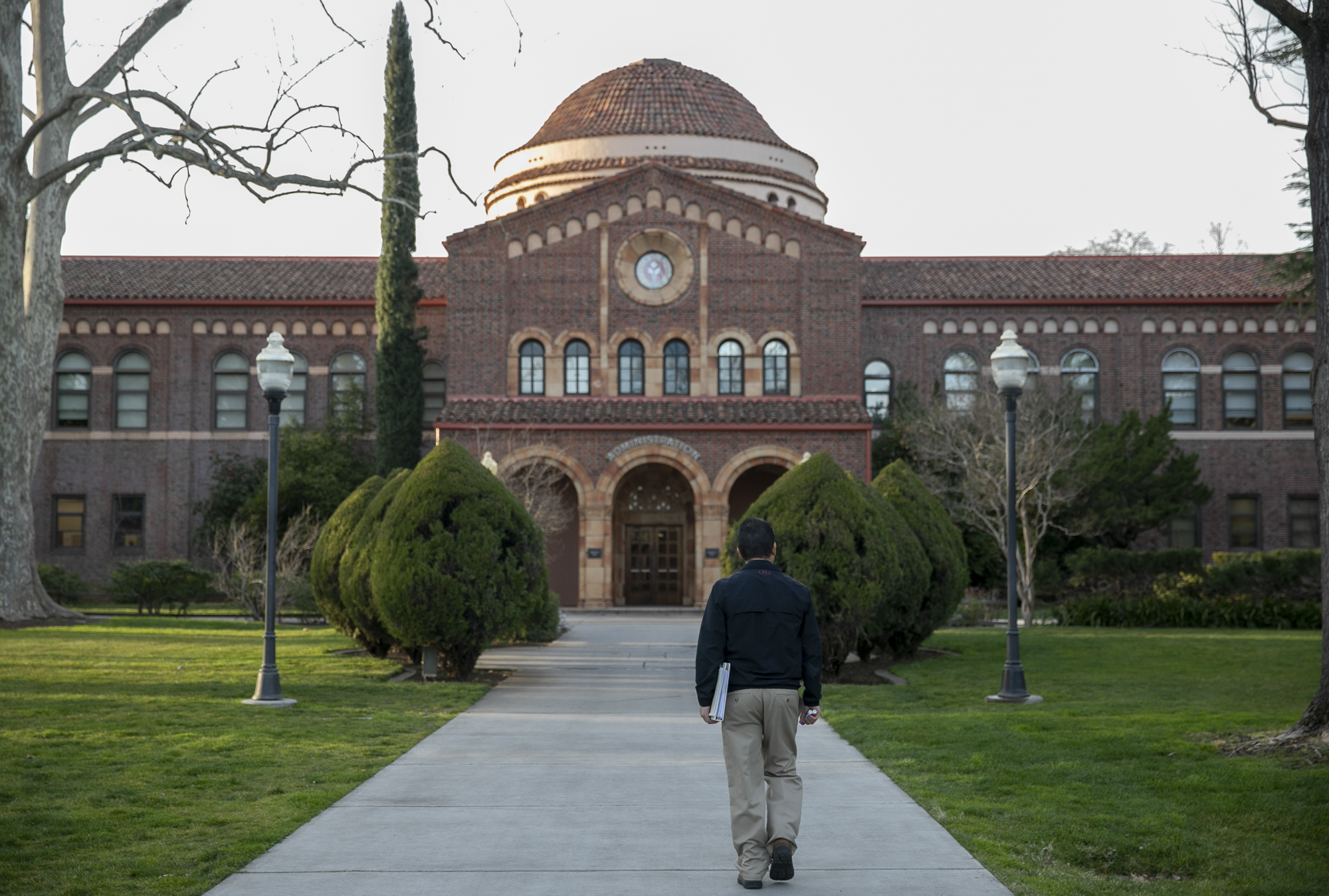 A person wearing a dark shirt and grey pants walks toward a building on a campus.