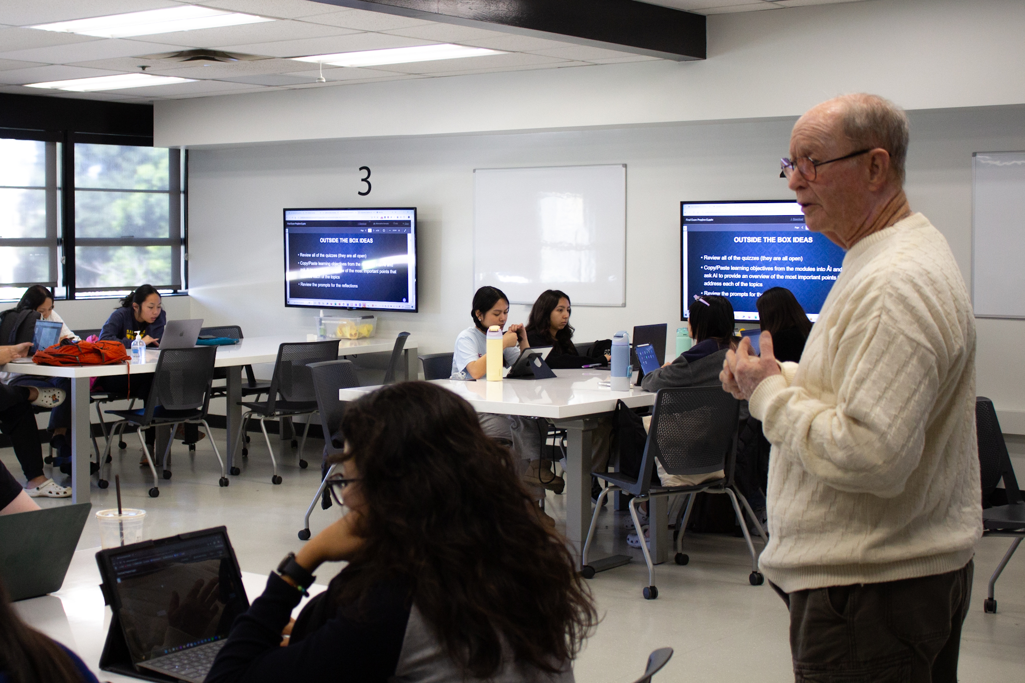A white man wearing glasses and a light colored sweater stands near seated students at desks with laptops and computer monitors on the walls in a classroom.