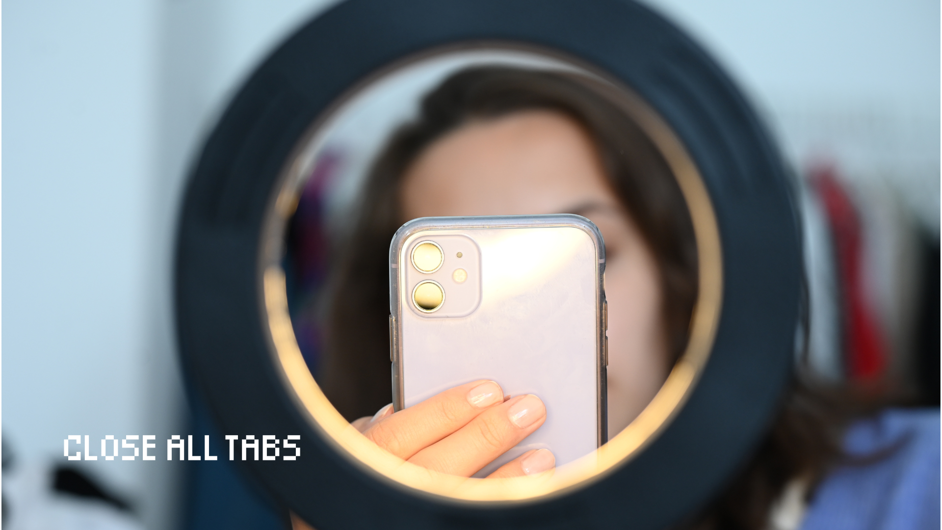 A woman with dark brown hair sits in front of a ring light, holding up a phone and obscuring her face. The phone and her hand are in focus, while the rest of the image is blurred. The words "Close All Tabs" appear in the bottom left hand side of the image.