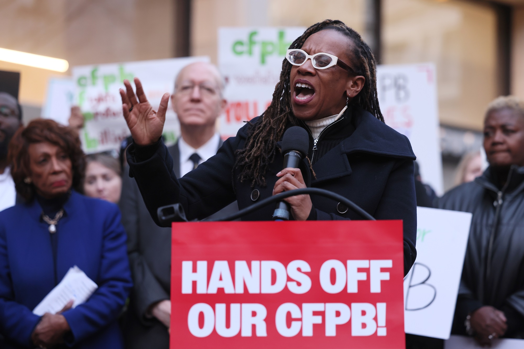A Black woman in glasses speak into a microphone at a rally, with people standing behind her. In front of her is a sign that says 'Hands off our CFPB!'