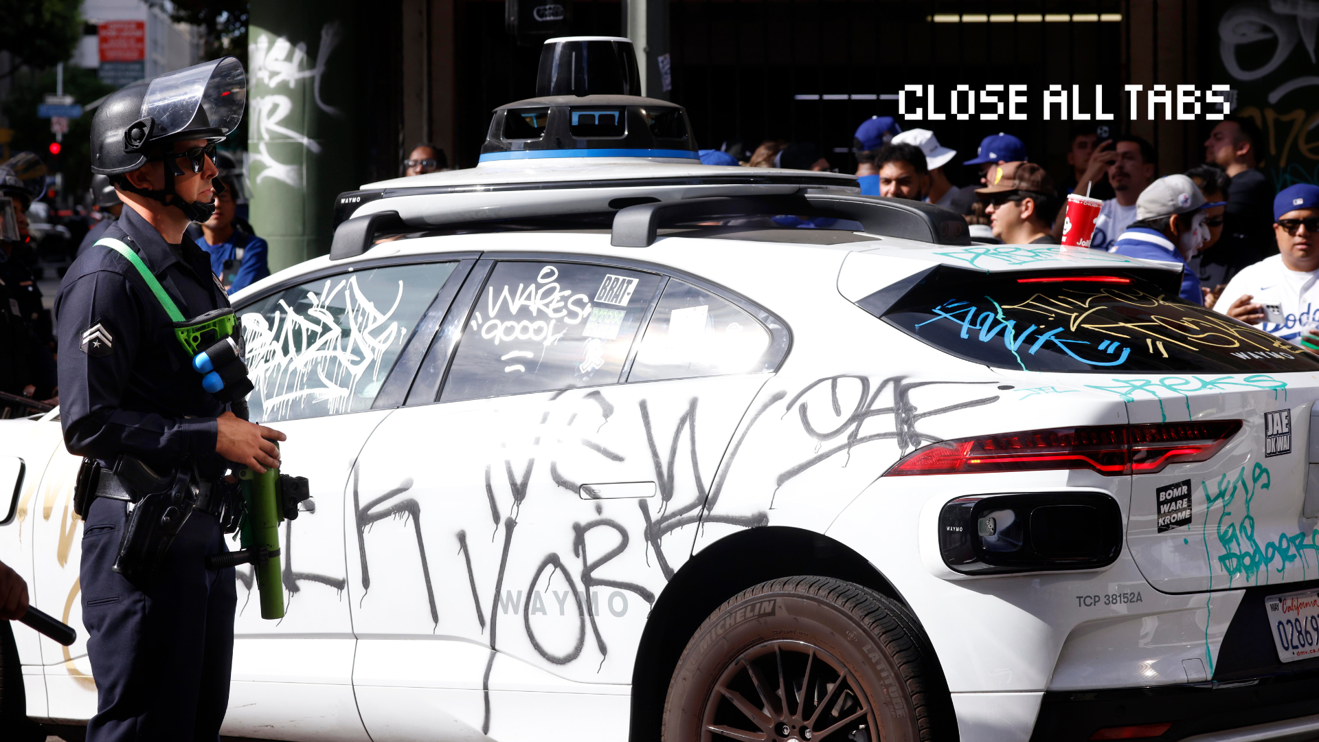 A man wearing a police uniform and a riot face shield stands next to a white Waymo vehicle that is covered in multicolor graffiti tags. A crowd of people wearing Dodgers paraphernalia surrounds the car.