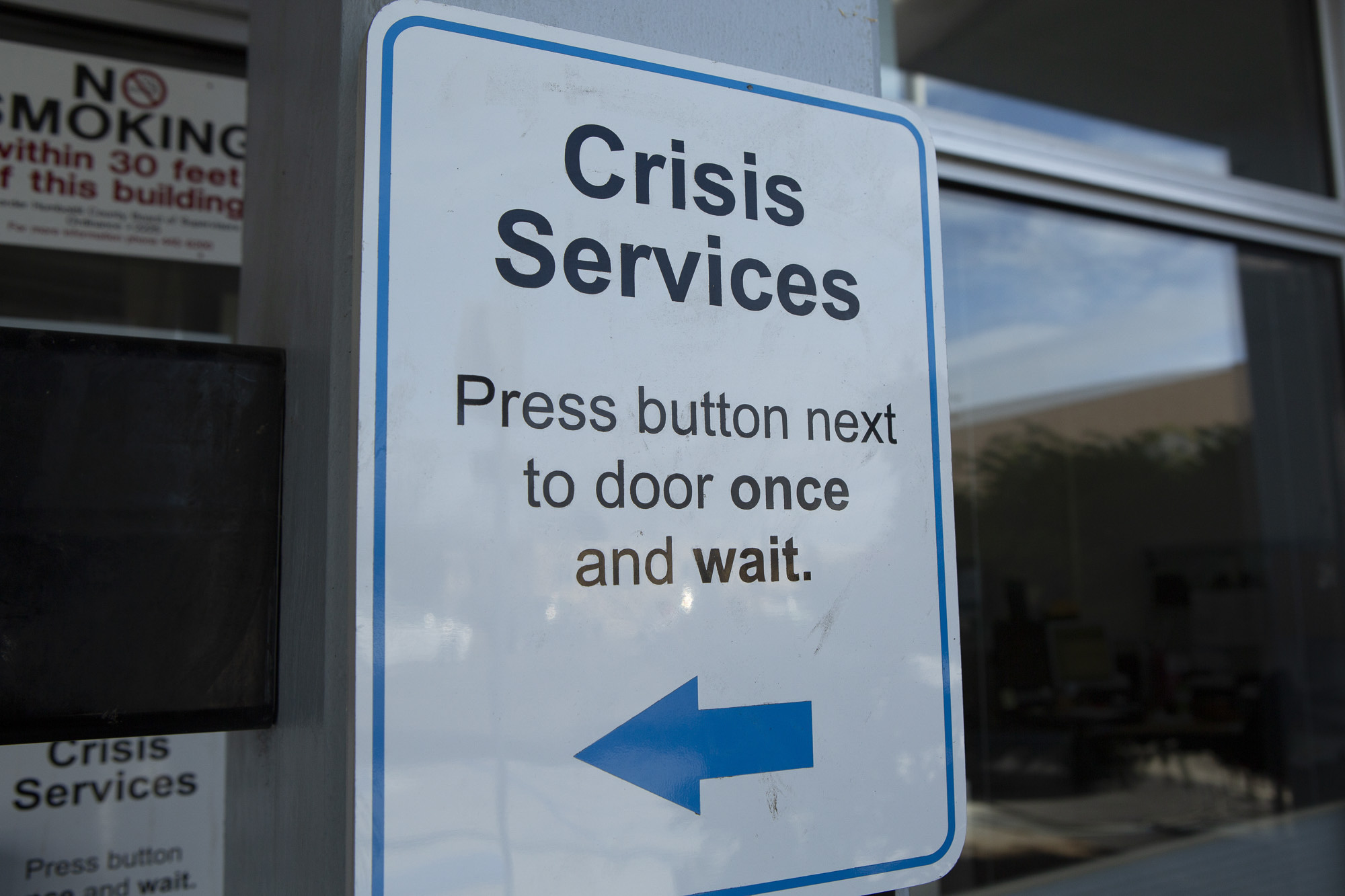 A white and blue sign that reads "Crisis Services Press button next to door once and wait." There is a blue arrow pointing right.