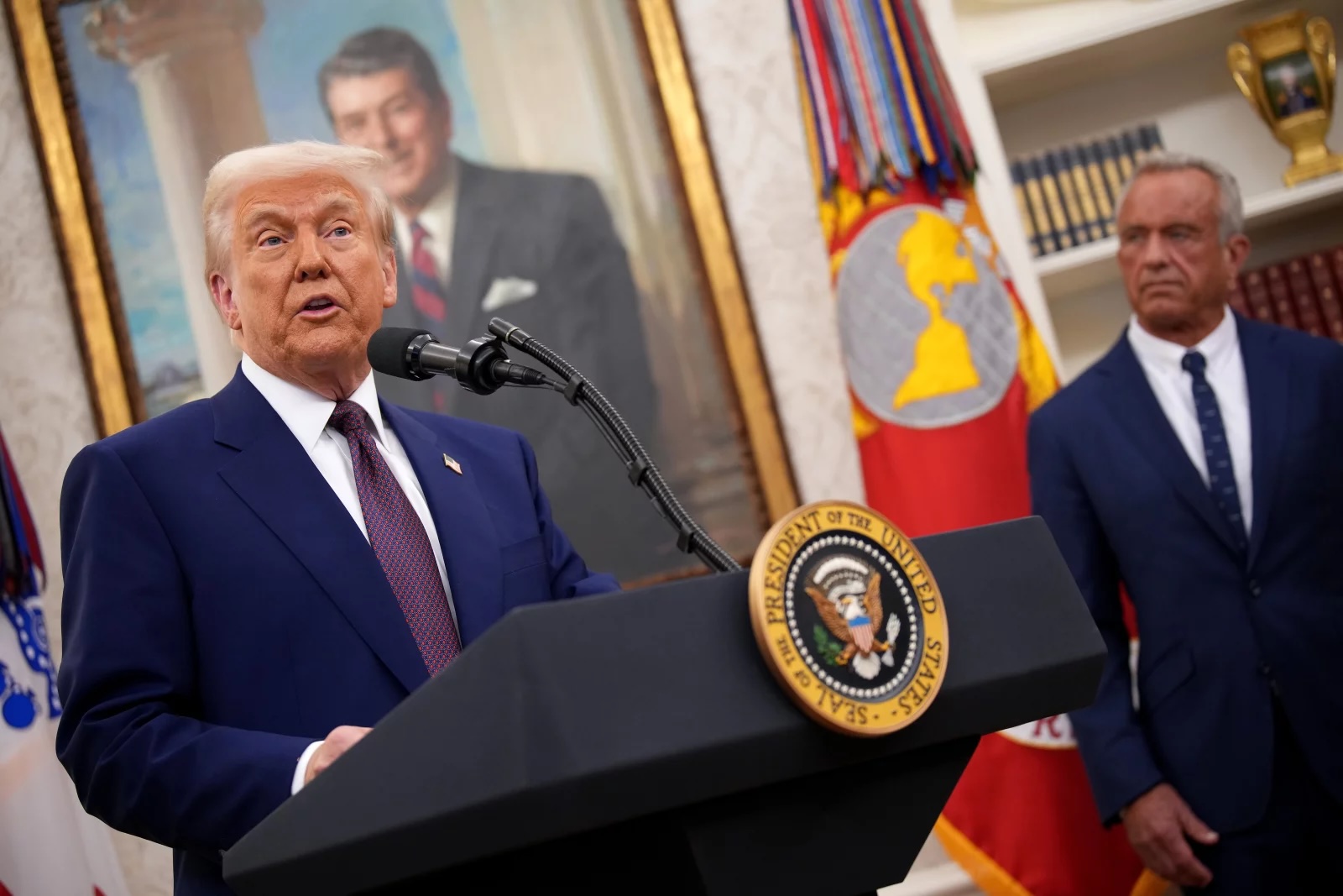 A solarium-tanned older and portly white man with a blue suit and tie speaks as another man in a blue suit and tie watches in an office, with a portrait of another older white man in a suit and tie hanging on the wall behind them.