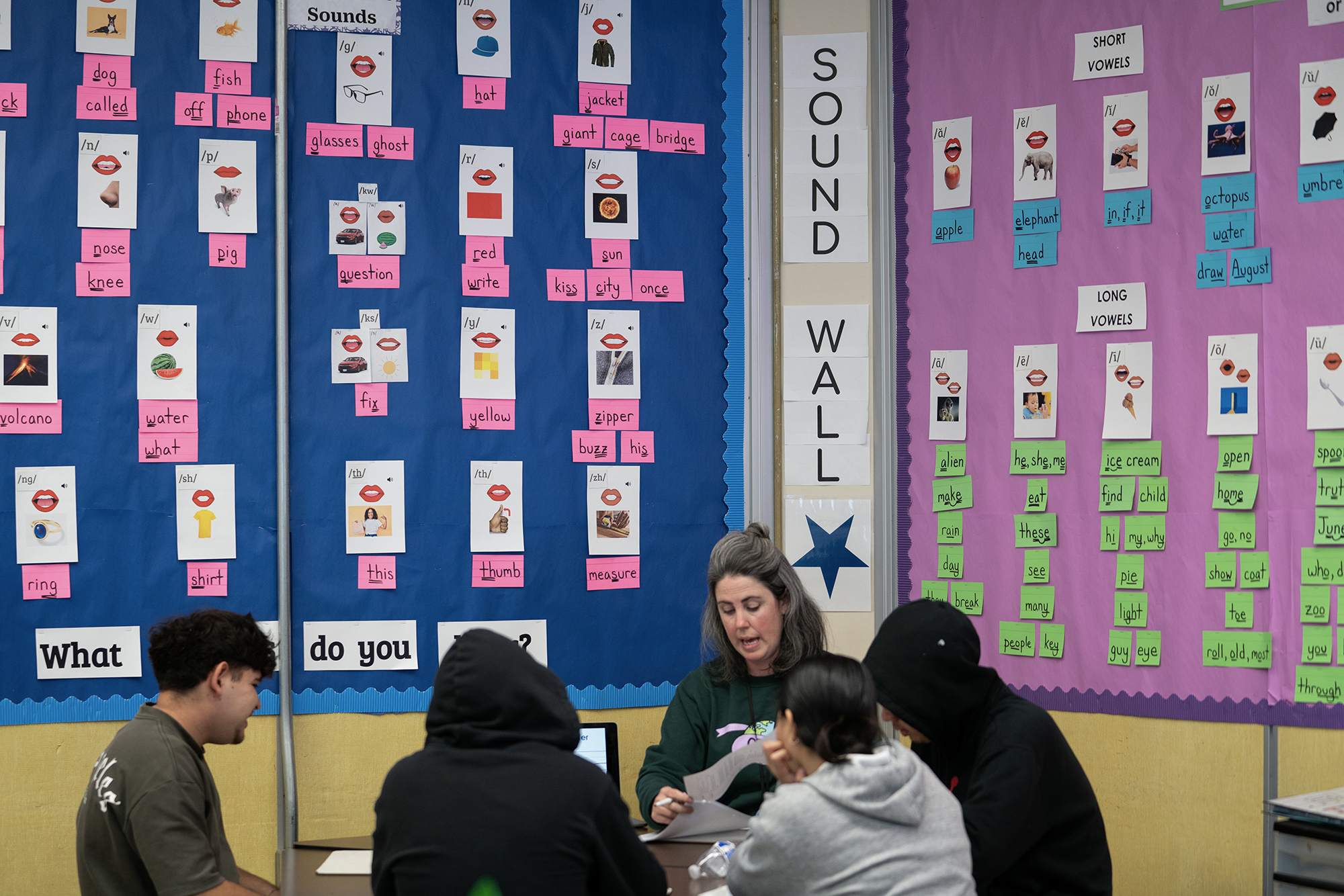 A white woman wearing a green sweater sits down at a desk surrounded by four students in a classroom.
