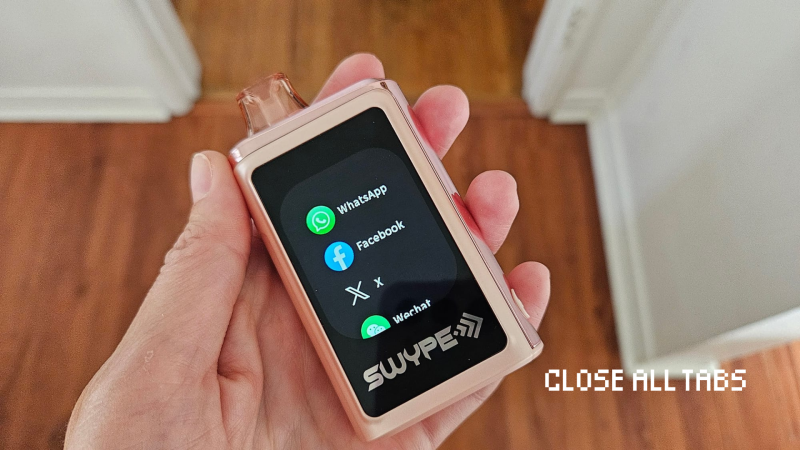 A pink SWYPE brand disposable vape is held in a person's left hand. The vape has a glossy touchscreen displaying icons for WhatsApp, Facebook, X (formerly Twitter), and WeChat. The background shows a reddish-brown wood floor and white door frames, slightly out of focus. In the lower right corner of the image, the phrase “CLOSE ALL TABS” appears in pixelated white font.