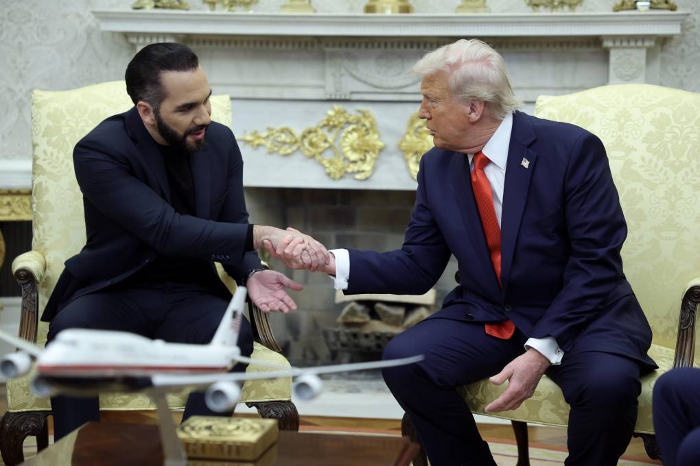 Two men wearing suits shake hands while seated in chairs.