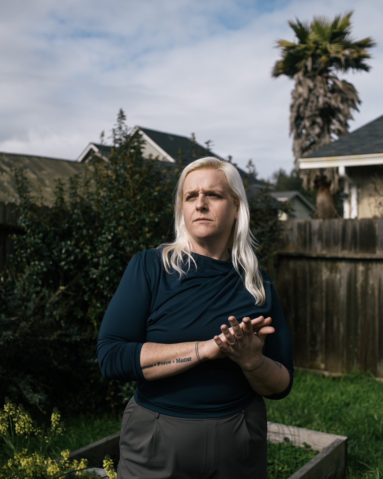 A white woman wearing a green shirt rubs her hands together and stands outside in a backyard.