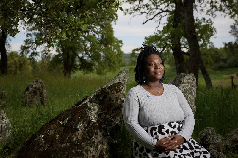 A Black woman wearing a white shirt and black and white skirt sits on a rock outside.
